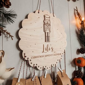 Può includere: Un calendario dell'Avvento in legno rotondo con bordi smerlati, caratterizzato da una figura di schiaccianoci incisa al laser, il testo 'Lili's CHRISTMAS ADVENT CALENDAR' e fori lungo il bordo inferiore per appendere sacchetti di carta.