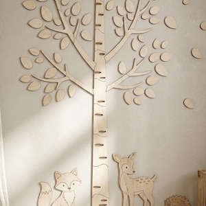 May include: Wooden wall decor featuring a birch tree with leaves, a fox, a deer, and a hedgehog. The tree trunk has measurement markings. The light-colored wood pieces are mounted on a neutral-toned wall, creating a woodland theme.
