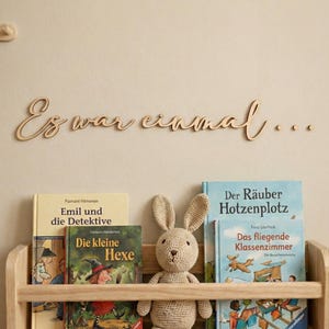 Può includere: Una libreria in legno con libri per bambini e un coniglietto di peluche. I libri includono titoli come "Emil e i detective" e "La piccola strega". Sopra lo scaffale, è visualizzata la frase "Es war einmal...".