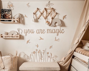 May include: A white wooden sign with the words "You are magic" in a whimsical font. The sign is decorated with small wooden cutouts of birds, flowers, and a rainbow. The sign is hanging on a white wall with a light brown background.