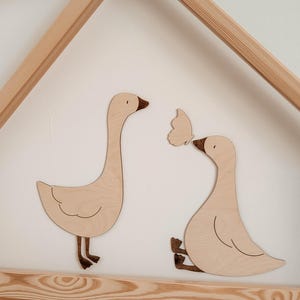 Op de afbeelding: Houten wanddecoratie met twee ganzen en een vlinder. De ganzen zijn lichtbruin met donkerbruine accenten, en de vlinder is lichtbruin. De kunst is gemonteerd op een witte muur, met een houten frame op de achtergrond.