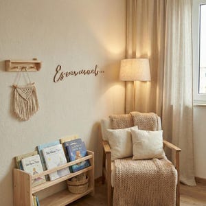 May include: A cozy reading nook with a wooden bookshelf filled with books, a macrame wall hanging, and a wooden chair with pillows and a knitted throw. The wall features the text "Es war einmal..." in cursive.