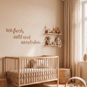 May include: A nursery room with a wooden crib, shelves, and a play gym. The walls are a neutral beige color, and the room is decorated with soft toys and a woven rug. The text "sei frech, wild und wunderbar" is on the wall.