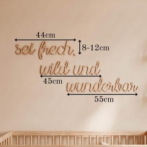May include: Wooden wall art in a cursive font that reads "sei frech, wild und wunderbar". The top word is 44cm wide, the middle word is 45cm wide, and the bottom word is 55cm wide. The wall art is displayed above a wooden crib.