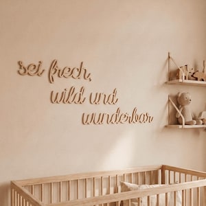 May include: Wooden wall art spelling out "sei frech, wild und wunderbar" in a nursery setting. The room features a wooden crib, shelves with toys, and a neutral color palette.