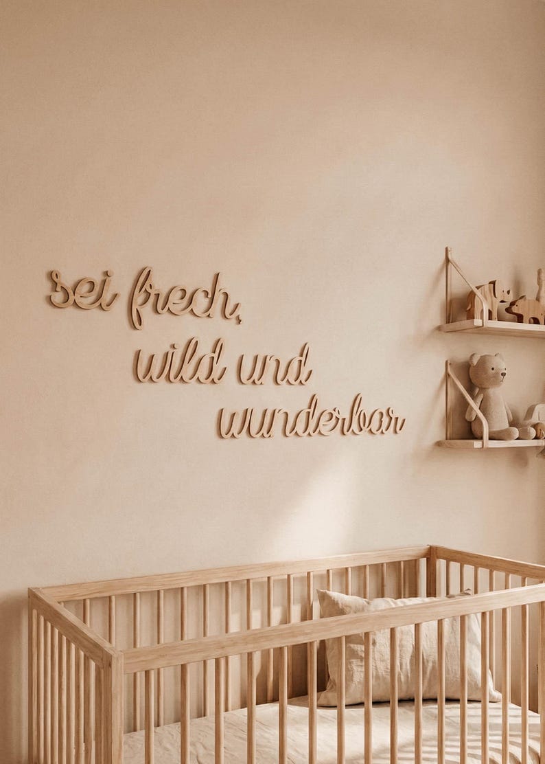 May include: Wooden wall art spelling out "sei frech, wild und wunderbar" in a nursery setting. The room features a wooden crib, shelves with toys, and a neutral color palette.