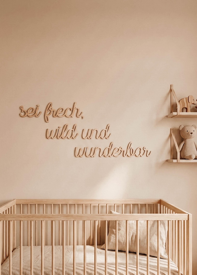 May include: Wooden wall decor with the German phrase "sei frech, wild und wunderbar" in a cursive font. The nursery scene includes a wooden crib, shelves with toys, and a neutral color palette.