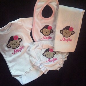 May include: A set of baby clothes featuring a monkey design with pink flowers and the name "Maylee" embroidered on each piece. The set includes a onesie, a bib, a burp cloth, and a pair of bloomers.