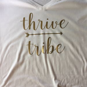 May include: White t-shirt with gold lettering that reads "thrive tribe" with an arrow pointing to the right.
