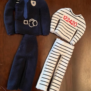 May include: Two elf-sized outfits: a blue police officer uniform with a badge, handcuffs, and a tie, and a white and black striped prisoner uniform with the number 122420.