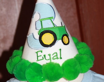 May include: A white birthday party hat with green pom-poms around the base. The hat has a blue number 3 and a green tractor embroidered on it. The name "Eyal" is embroidered below the tractor.