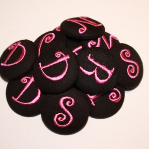 May include: A pile of black fabric buttons with pink embroidered letters. The letters include D, S, and N.
