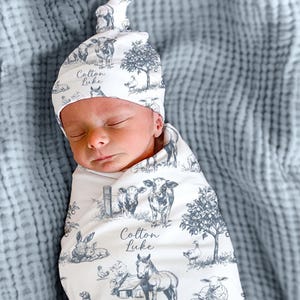 A sleeping newborn baby wrapped in a blanket with a pattern of farm animals, including a cow, horse, and sheep, along with the name "Colton Luke" printed on the blanket.