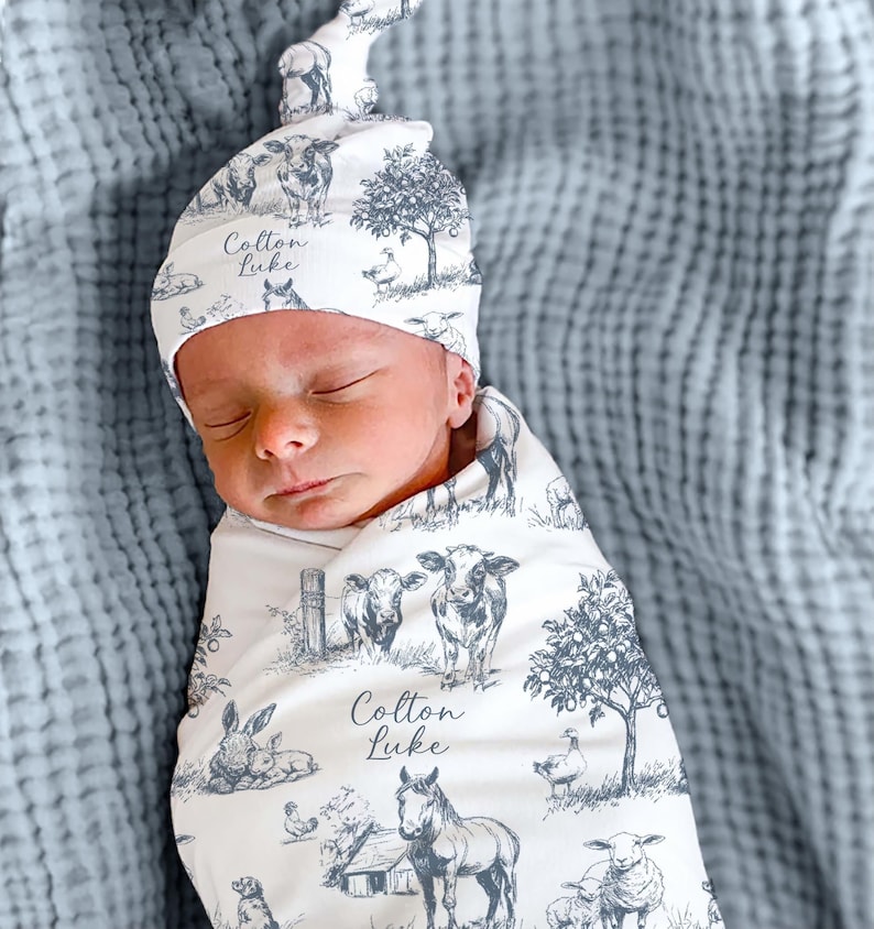 A sleeping newborn baby wrapped in a blanket with a pattern of farm animals, including a cow, horse, and sheep, along with the name "Colton Luke" printed on the blanket.