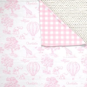 May include: A baby blanket with a pink and white toile pattern featuring giraffes, elephants, hot air balloons, and trees. The reverse side has a pink and white gingham pattern. The name "Charlotte" is printed on the blanket.