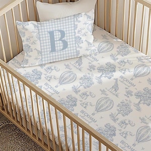 May include: A baby crib with a blue and white patterned bedding set. The set includes a pillow with a large blue letter "B" on a gingham background, and a fitted sheet with a repeating pattern of hot air balloons, giraffes, and elephants. The name "Beckham Edward" is printed on the sheet.