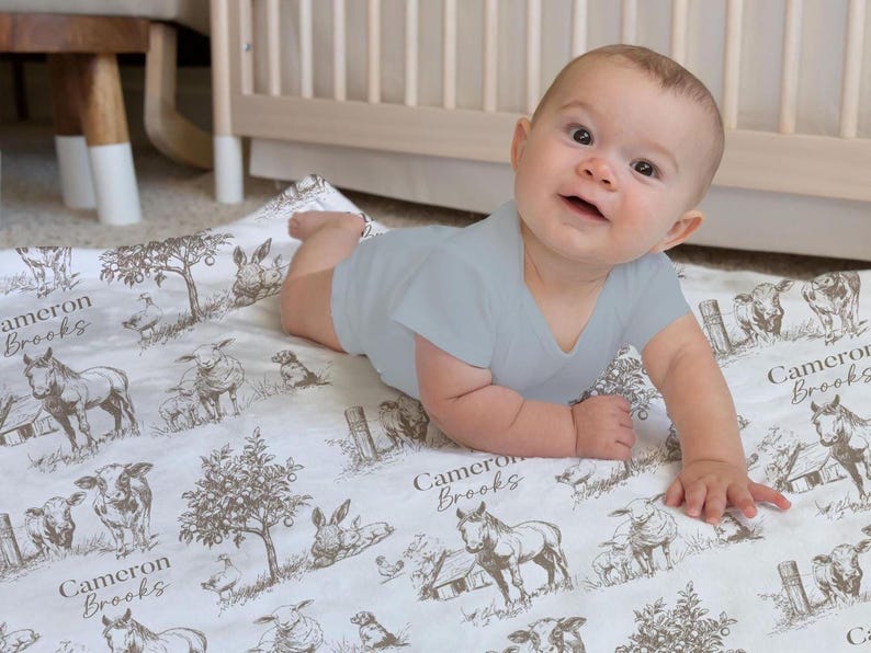 A baby is lying on a blanket with a farm animal print, smiling at the camera.