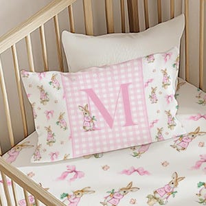 May include: A crib set featuring a pillow and fitted sheet with a bunny theme. The pillow has a pink gingham center with a large pink letter "M". The sheet and pillow have a white background with pink bunnies and bows.
