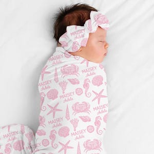 May include: A baby swaddled in a white blanket and wearing a matching headband, both adorned with a pink sea-themed pattern. The pattern includes starfish, seahorses, shells, and the name "MAISEY Adele".