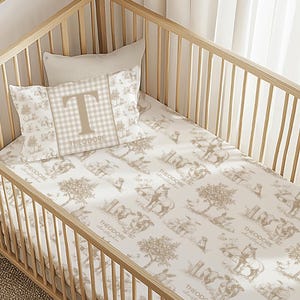 May include: A light wood crib with bedding featuring a toile pattern in beige and white. The bedding includes a pillow with a large letter "T" and the name "Theodore". The pattern depicts scenes with horses and trees.
