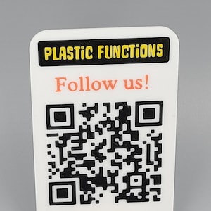 May include: A white plastic sign featuring a QR code and the text "Follow us!". The top of the sign has the words "PLASTIC FUNCTIONS" in yellow letters on a black background. The sign is supported by a white stand.
