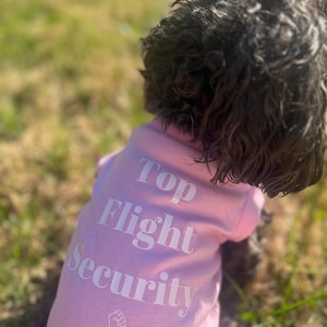 May include: A pink dog t-shirt with the words "Top Flight Security" in white block letters. The shirt also features a graphic of a fist. The dog has dark, curly fur and is seen from the back.