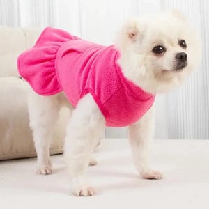 May include: A small white dog wearing a bright pink fleece dress. The dress has a ruffled skirt and a fitted bodice. The dog is standing on a white surface, with a neutral background.
