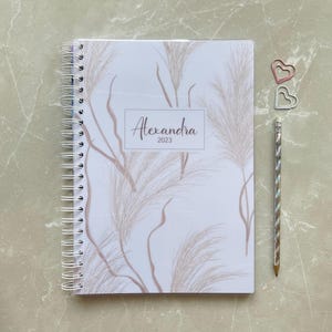 Peut inclure: Un agenda blanc avec une reliure spirale et un motif floral. L'agenda porte le nom "Alexandra 2023" imprimé sur la couverture.