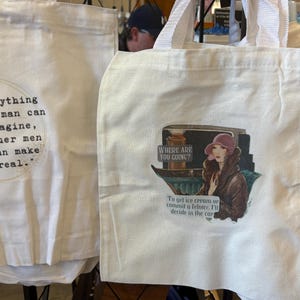 May include: Two white tote bags with different designs. The first bag has a quote about imagination printed on it. The second bag has a vintage illustration of a woman in a car with the text "Where are you going? To get ice cream or commit a felony. I'll decide in the car."
