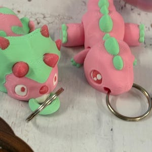 May include: Two pink and green dinosaur keychains with silver key rings. The dinosaurs are 3D printed and have a cartoonish style.