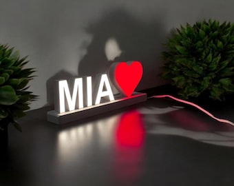 May include: A white LED light-up sign that spells "MIA" with a red heart symbol. The letters and heart are illuminated, casting a warm glow. The sign is on a dark surface, with a green plant in the background.