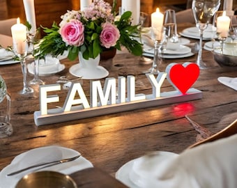 May include: White illuminated sign with the word "FAMILY" in block letters and a red heart. The sign is on a wooden table with candles and flowers.