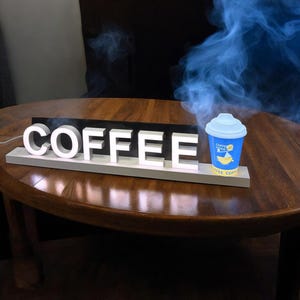 Könnte beinhalten: Ein weißes beleuchtetes Schild mit dem Wort "COFFEE" in großen Buchstaben. Eine kleine blaue und gelbe Kaffeetasse mit aufsteigendem Rauch steht auf dem Schild.