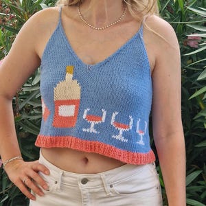 Peut inclure: Un débardeur en tricot bleu clair avec une bouteille de vin rose et quatre verres à vin blancs brodés sur le devant. Le débardeur a une bordure rose corail.