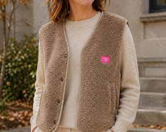 Könnte beinhalten: Eine ärmellose braune Sherpa-Fleece-Weste mit V-Ausschnitt, Knopfleiste auf der Vorderseite und einem kleinen rechteckigen rosa Aufnäher auf der linken Brust mit dem Text LOVELY DAY, getragen über einem cremefarbenen langärmeligen Strickpullover.