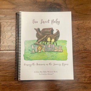 May include: A white book with a brown and black illustration of a boat with animals on it. The title of the book is "Our Sweet Baby" and the subtitle is "Keeping the Memories as the Journey Begins". The book is a "Glory Bee Baby Memory Book" by Carolyn Altman.