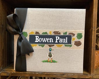 May include: A personalized photo album with the name "Bowen Paul" on a black label. The album is wrapped with a black ribbon and features a duck illustration. The background is a neutral linen texture.