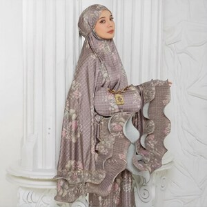 May include: A mauve-colored prayer garment set with a floral pattern, including a head covering and a matching handbag. The set features ruffled edges and a decorative clasp on the handbag.