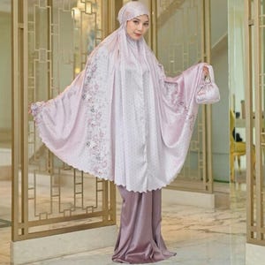 May include: A light purple prayer garment set, including a head covering, shawl, and matching purse. The set features a floral pattern and a scalloped edge. The woman is wearing a matching skirt.