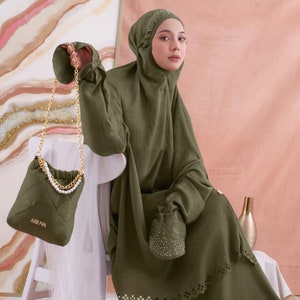 May include: A woman wearing a green prayer dress with a matching head covering and a small green quilted bag with a gold chain. The bag has the word "AIBUNA" printed on it.