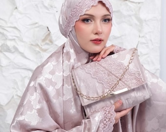 May include: A dusty rose satin prayer garment featuring a matching head covering with intricate lace trim. The fabric displays a subtle tonal floral pattern. A coordinating rectangular handbag with a gold chain strap and lace detail is included.