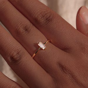 May include: A gold ring featuring a rectangular, clear gemstone. The ring is on a person's finger, with the hand and part of a light-coloured fabric visible in the background. The ring's band is thin and delicate.