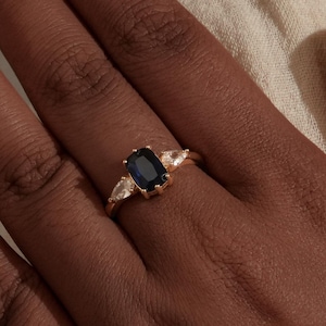 May include: A gold ring with a dark blue, rectangular central gemstone and two smaller, triangular, white stones. The ring is displayed on a hand, set against a light background.