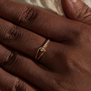 May include: A gold ring with a distinctive design, featuring a row of small, clear stones along the top band. The ring has an open shape with a pointed element. The ring is worn on a hand with dark skin, against a neutral background.