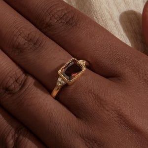 May include: A gold ring featuring a rectangular, deep red gemstone and two smaller white stones. The ring is on a hand, with a cream-coloured fabric in the background. The ring has a detailed, textured design around the gemstones.