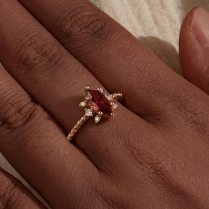 Peut inclure: Une bague en or ornée d'une pierre précieuse rouge foncé en forme de poire en son centre. La bague est ornée de plus petites pierres précieuses transparentes et de détails délicats. La bague est sur la main d'une personne.