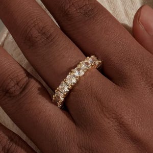 May include: A gold ring set with multiple clear gemstones, worn on a hand. The ring's band is gold, and the gemstones are evenly spaced. The hand has a medium skin tone, and the background is a neutral colour.