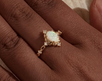 May include: A gold ring with an oval, white opal center stone. The ring has a decorative, filigree design with small, clear accent stones. The ring is on a person's hand, with a neutral background.