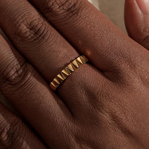 May include: A gold-colored ring with a textured, geometric design. The ring is worn on a hand with dark skin, and the background is a soft, neutral fabric. The ring's design features a series of small, pointed shapes.