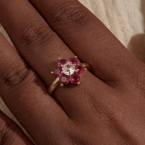 Peut inclure: Une bague en or avec un motif floral, ornée d'une pierre transparente centrale entourée de pierres rouges. La bague est présentée sur une main à la peau foncée, soulignant le contraste des couleurs. L'anneau de la bague est en or poli.